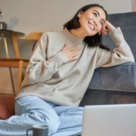 Portrait of dreamy woman sitting with laptop on floor, watching on computer and drinking coffee, enjoying cozy days at home.